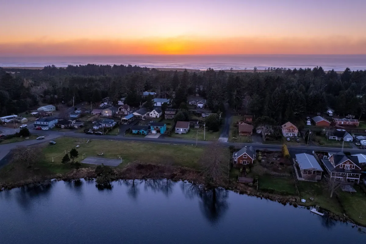 A breathtaking view of the sun setting over the ocean, with houses lining the shoreline, a reflective water surface, and a lush green park area in the foreground. The image captures families and friends walking along the shore, picnicking in the park, and casting fishing lines into the water, all set against the tranquil glow of dusk.