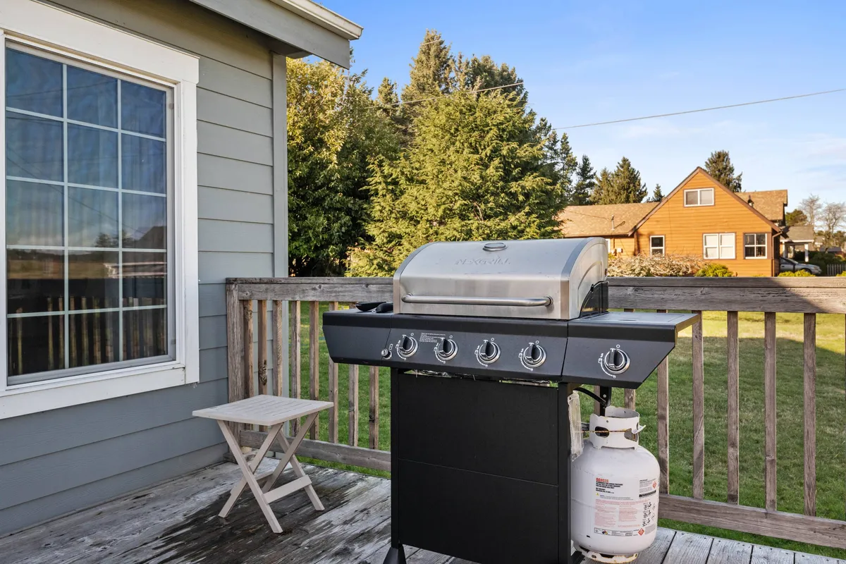 A spacious wooden deck equipped with a grill, surrounded by large windows offering views into the home and out over natural scenery. The deck is set up for outdoor gatherings, with the promise of sunset dinners, celebrations, and relaxed afternoons overlooking the landscape.