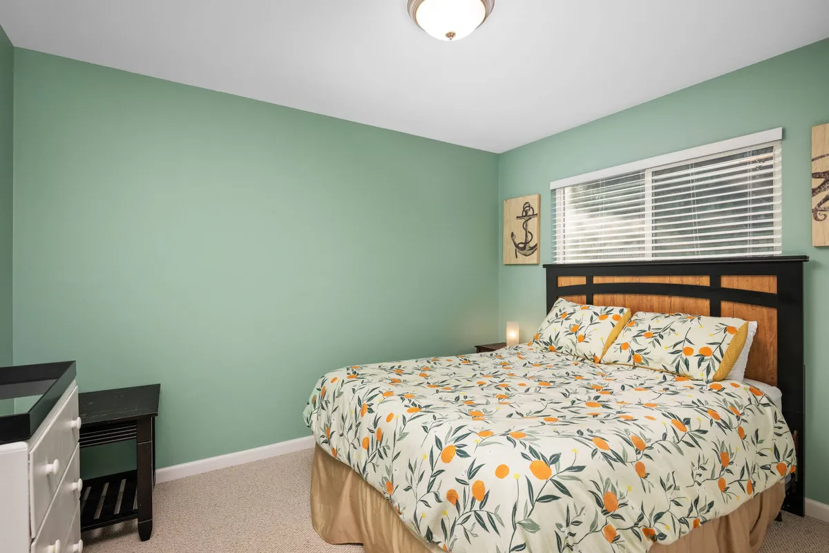 A serene bedroom featuring a bed with floral bedding against green walls, a window with blinds letting in natural light, and a cozy atmosphere ideal for restful sleep. The room includes a nightstand and soft lamp lighting, creating a tranquil retreat.