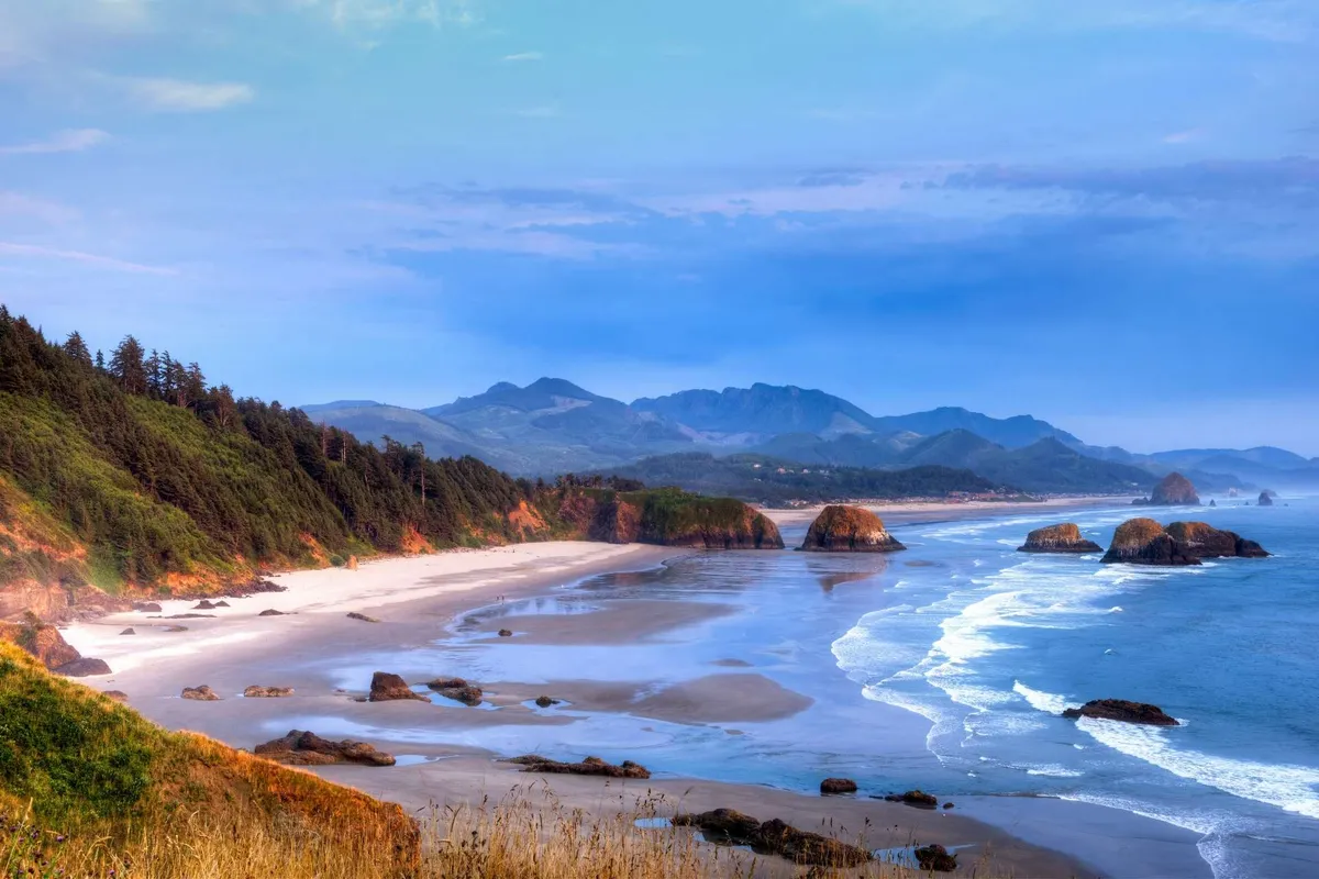 This image captures a broad sandy beach with distant mountains and unique rock formations emerging from the water. The scene is tranquil and inviting, perfect for activities like beachcombing, photography, and hiking nearby trails. The soft light suggests the possibility of a sunset, adding to the serene ambiance.