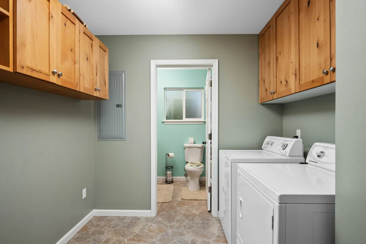 A practical interior image of a laundry area featuring modern washing machines, wooden cabinets for storage, and a glimpse of an adjoining room. The space is organized and ready for post-adventure clean-ups.