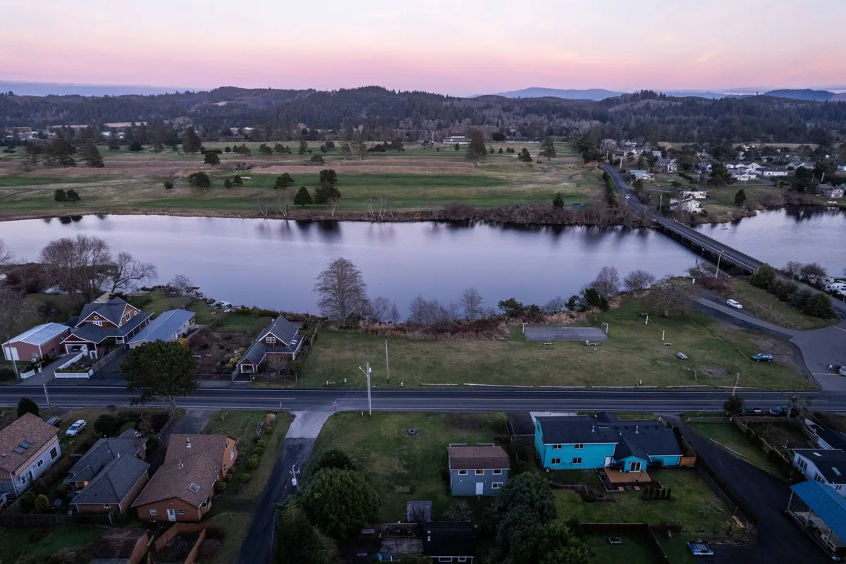 A wide view over the river, houses, a bridge, and green fields, inviting activities like fishing, biking, and picnicking. The tranquil river scene offers context for the neighborhood's access to outdoor recreation and scenic beauty close to parks and beaches.